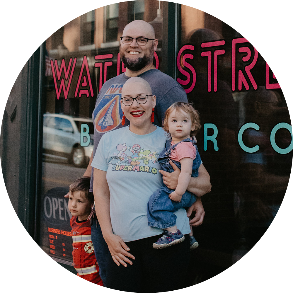 A young family of four smiles at the camera, standing in front of the Water Street Barber Co. Julia, the mother, is entirely bald and holds one of the two children. The older child stands next to their father, who is also bald like Julia.
